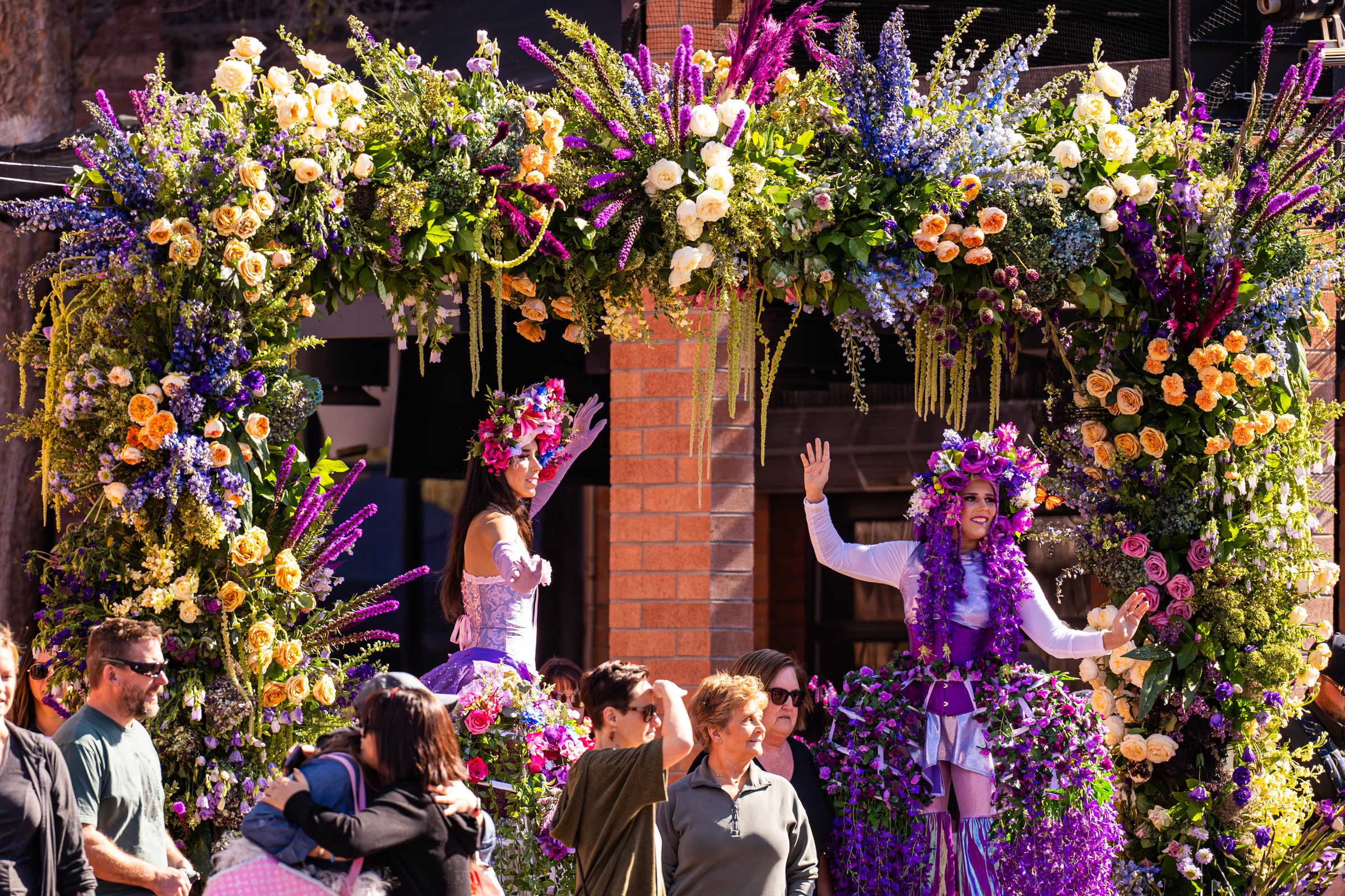 Vibrant floral archway with performers in colorful costumes celebrating Tempe Blooms festival, surrounded by attendees enjoying the festive atmosphere in Downtown Tempe, AZ.
